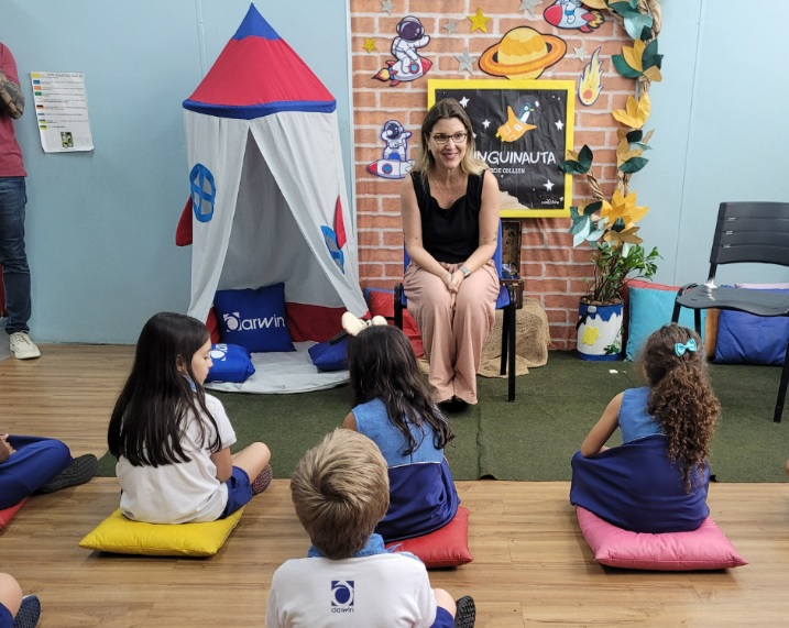 Mulher loira, sentada em cadeira, falando com crianças em uma escola.