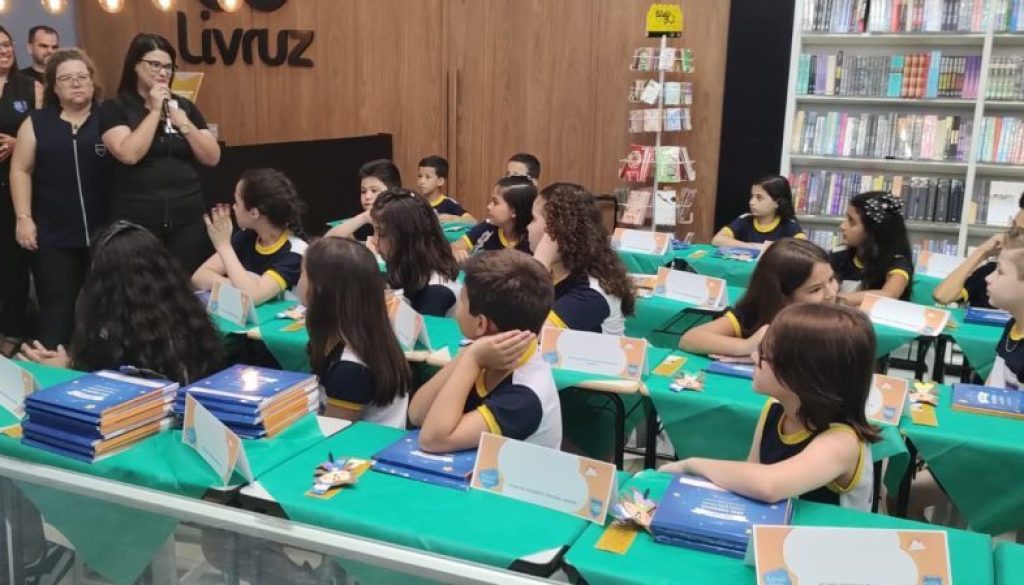 Professoras em pé olhando para alunos sentados na cadeira, apoiando-se em uma mesa com livros.