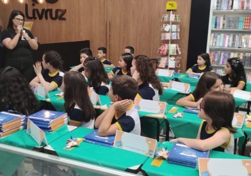 Professoras em pé olhando para alunos sentados na cadeira, apoiando-se em uma mesa com livros.