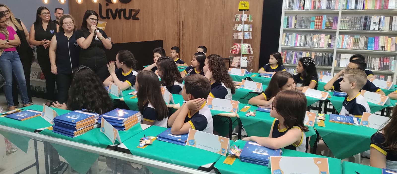 Professoras em pé olhando para alunos sentados na cadeira, apoiando-se em uma mesa com livros.
