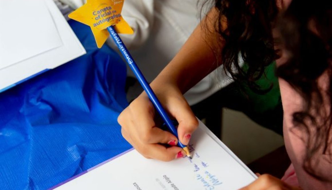 Menina de pele branca, com os cabelos castanho escuro e compridos, fazendo a leitura da frente de sua arte, seu livro, enquanto escreve seu nome com um lápis azul, com uma estrela na ponta escrito Estante Mágica.