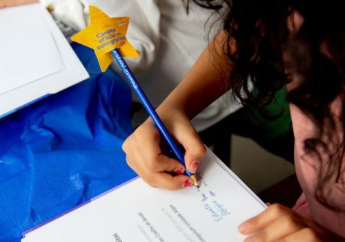 Menina de pele branca, com os cabelos castanho escuro e compridos, fazendo a leitura da frente de sua arte, seu livro, enquanto escreve seu nome com um lápis azul, com uma estrela na ponta escrito Estante Mágica.