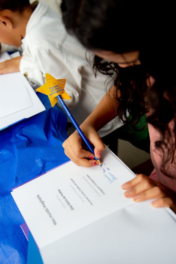 Menina de pele branca, com os cabelos castanho escuro e compridos, fazendo a leitura da frente de sua arte, seu livro, enquanto escreve seu nome com um lápis azul, com uma estrela na ponta escrito Estante Mágica.