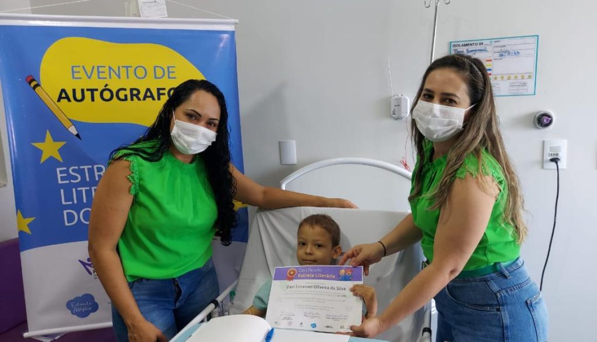 Duas professoras com máscara posando ao lado de uma criança deitada em uma cama de hospital, em tratamento contra o câncer infantil. Ao fundo, banner sobre Evento de Autógrafos.