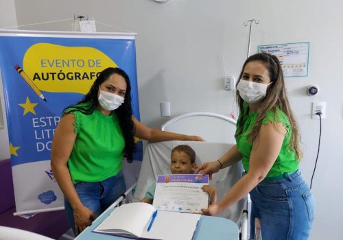 Duas professoras com máscara posando ao lado de uma criança deitada em uma cama de hospital, em tratamento contra o câncer infantil. Ao fundo, banner sobre Evento de Autógrafos.