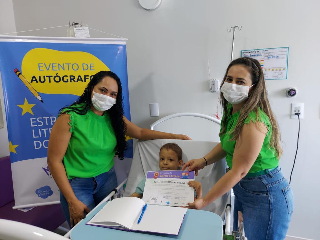 Duas professoras com máscara posando ao lado de uma criança deitada em uma cama de hospital, em tratamento contra o câncer infantil. Ao fundo, banner sobre Evento de Autógrafos.