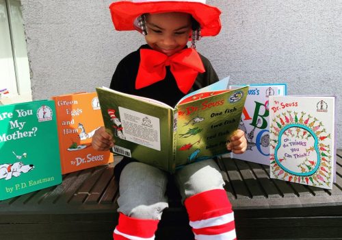 Menina de etnia negra, aparentando ter 3/4 anos de idade, com um chapéu grande, listrado de vermelho e branco sobre sua cabeça, lendo um livro com título em inglês, chamado: “One fish, two fish, three fish, four fish”, do Dr. Seuss, para mostrar os efeitos de uma educação antirracista.