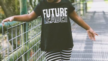 Criança negra de 4 / 5 anos, de pé em uma ponte, usando uma camiseta preta com a frase “Future Leader” estampada e calça legging listrada de preto e branco, e tênis preto e branco. Imagem destacada para o Mês da Consciência Negra.