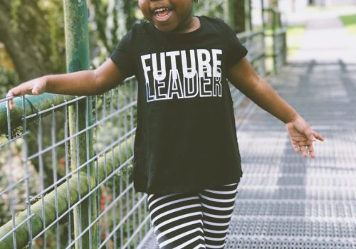 Criança negra de 4 / 5 anos, de pé em uma ponte, usando uma camiseta preta com a frase “Future Leader” estampada e calça legging listrada de preto e branco, e tênis preto e branco. Imagem destacada para o Mês da Consciência Negra.