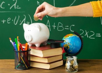 Imagem mostra quadro negro, mão com moeda e um porquinho sobre livros simbolizando educação financeira na escola.