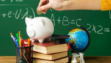 Imagem mostra quadro negro, mão com moeda e um porquinho sobre livros simbolizando educação financeira na escola.