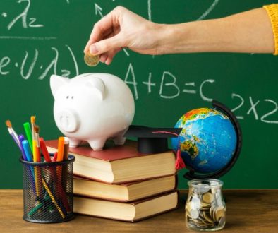 Imagem mostra quadro negro, mão com moeda e um porquinho sobre livros simbolizando educação financeira na escola.