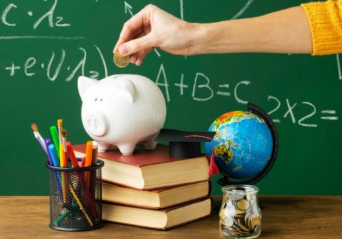 Imagem mostra quadro negro, mão com moeda e um porquinho sobre livros simbolizando educação financeira na escola.